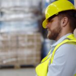 Exhausted young cargo worker sleeping leaning on large container in warehouse. Tired warehouse manager sitting on floor near cardboard boxes and napping during break