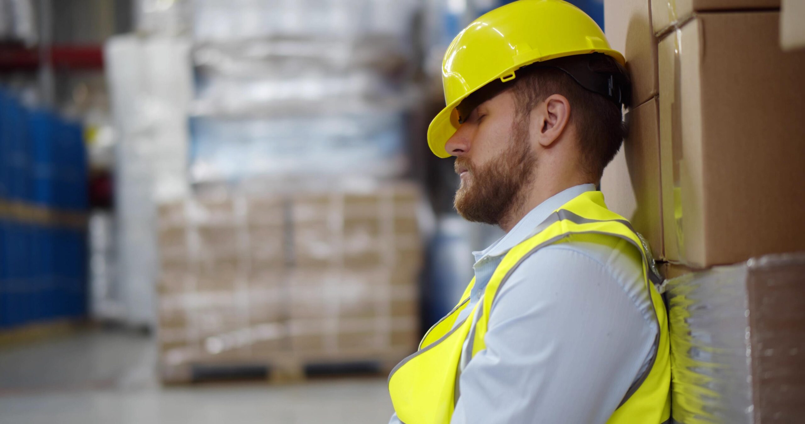 Exhausted young cargo worker sleeping leaning on large container in warehouse. Tired warehouse manager sitting on floor near cardboard boxes and napping during break