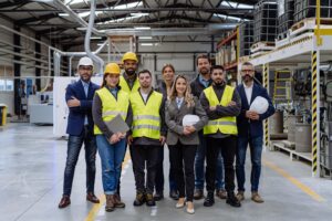 Full team of warehouse employees standing in warehouse. Team of workers, managers and female director in modern industrial factory, heavy industry, manufacturer. Group portrait.
